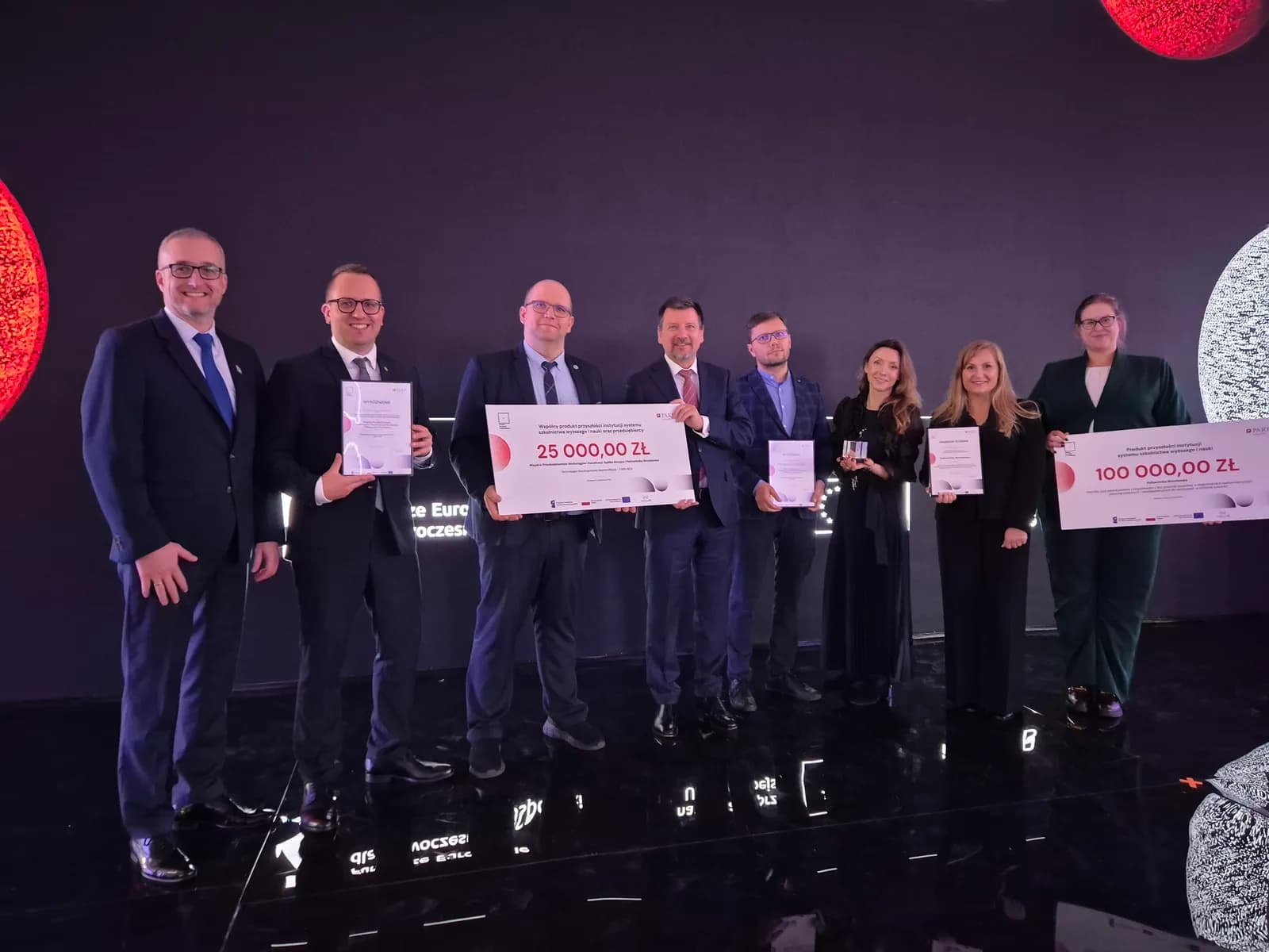 Eight people, five men and three women, stand on a stage holding certificates and large checks, posing for a group photo.