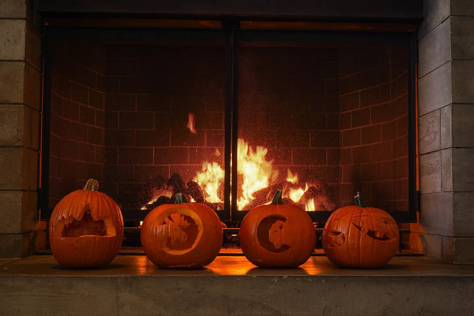 Four carved pumpkins sit on a hearth in front of a fireplace with a roaring fire.