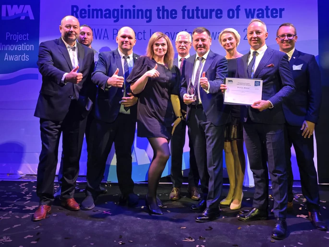 A group of ten people, six men and four women, stand on a stage with a blue background displaying "Reimagining the future of water," holding awards and giving thumbs up, with confetti on the floor.