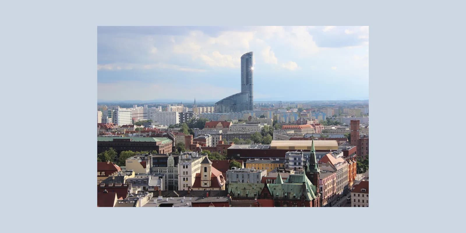 An aerial view of a sprawling cityscape with a distinct, curved skyscraper rising prominently above other buildings under a partly cloudy sky.