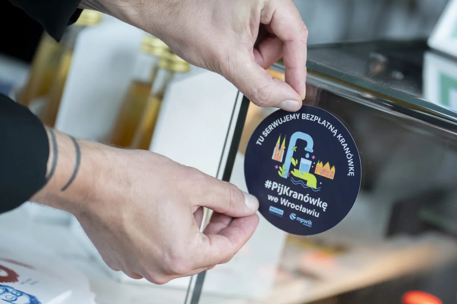 A person's hands are holding a circular dark blue sticker that says "TU SERWUJEMY BEZPŁATNĄ KRANÓWKĘ #PijKranówkę we Wrocławiu" with a graphic of a faucet, hands, and city skyline.