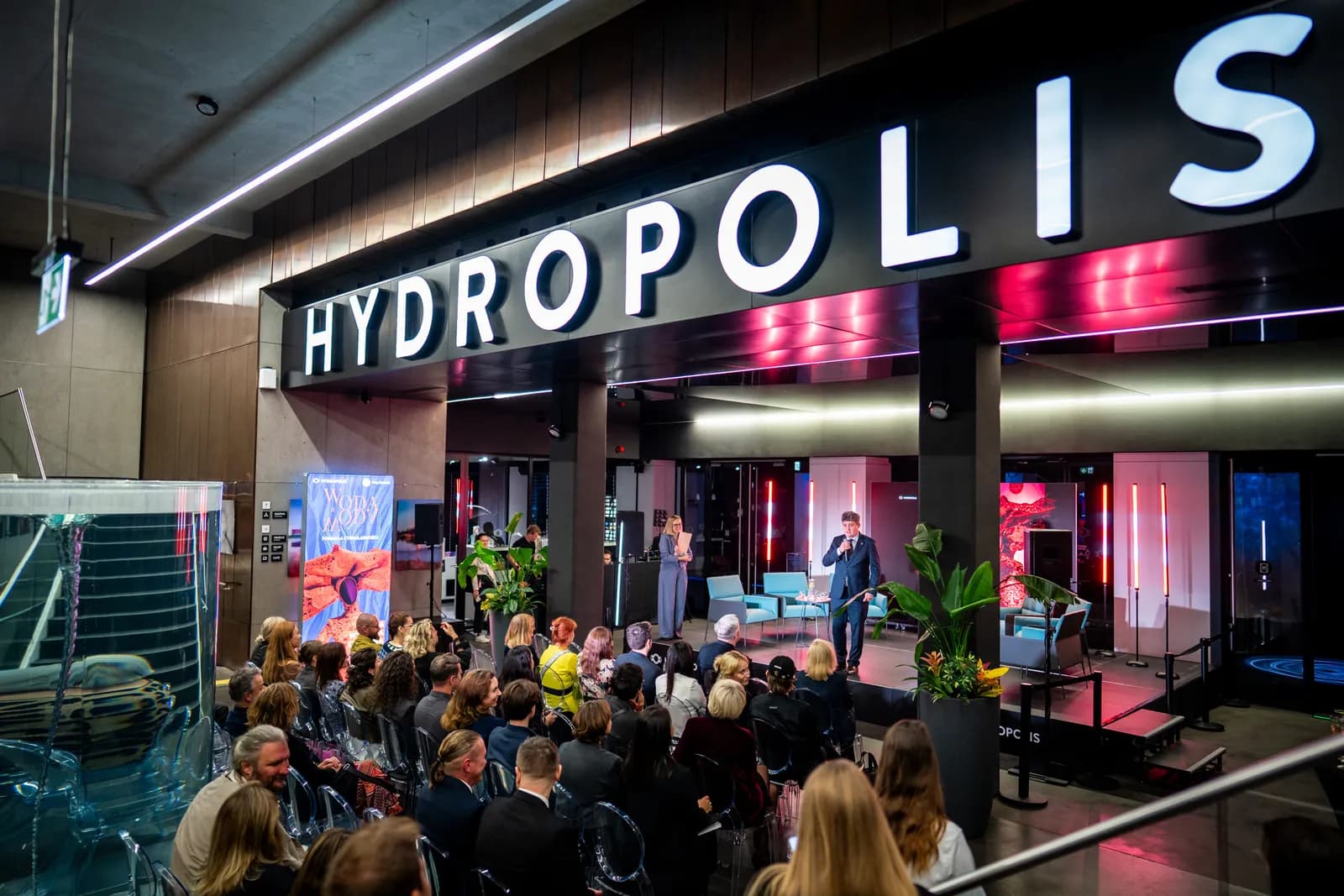 A man in a suit speaks into a microphone on a stage to an audience seated in a modern venue with a large "HYDROPOLIS" sign above him.